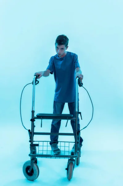 Πλήρης εικόνα ενός έφηβου ανάπηρου αγοριού με εγκεφαλική παράλυση που κοιτάζει την κάμερα, κάνοντας βήματα με τον περιπατητή του απομονωμένο σε μπλε φόντο με νέον φως. — Φωτογραφία Αρχείου