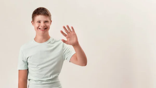 Menino deficiente feliz com síndrome de Down sorrindo e acenando para a câmera enquanto posando isolado sobre fundo branco — Fotografia de Stock