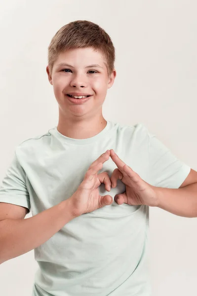 Joyful disabled boy with Down syndrome smiling at camera, making heart shape with his hands while standing isolated over white background — Photo