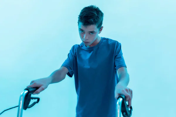 Portrait of teenaged disabled boy with cerebral palsy looking at camera, taking steps with his walker isolated over blue background with neon light — Stock Photo, Image