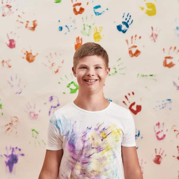 Alegre niño discapacitado con síndrome de Down sonriendo a la cámara mientras está de pie frente a la pared con muchas impresiones coloridas de las manos —  Fotos de Stock