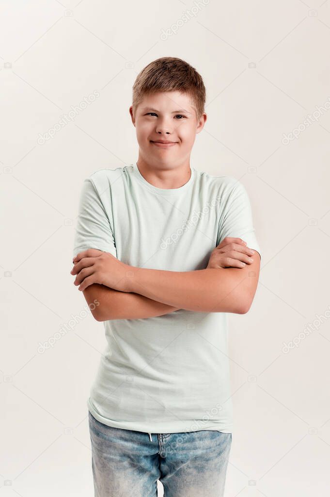 Teenaged disabled boy with Down syndrome smiling at camera while posing, standing with arms crossed isolated over white background