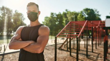 Maske takan, kameraya bakan, dışarıda kollarını kavuşturmuş, dışarıda spor yapan genç bir adam.