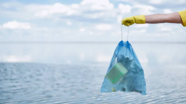 Voluntarios mano en guante de goma amarilla sosteniendo bolsa de basura con residuos de plástico y otra basura contra el mar y el cielo mientras limpian la playa — Foto de Stock