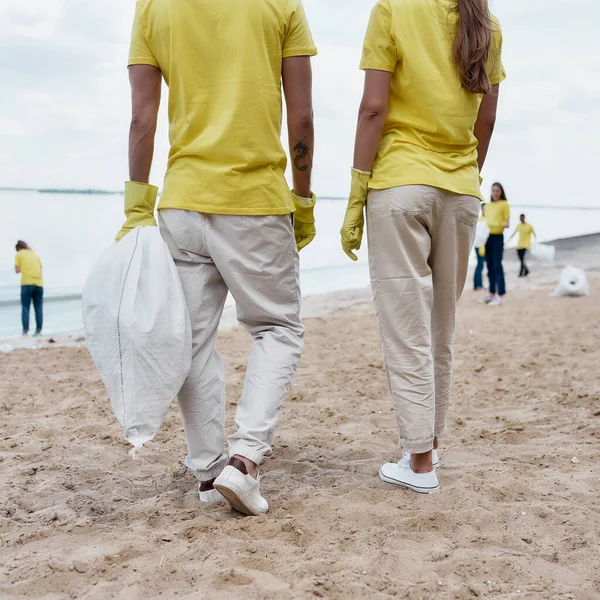 Vista posterior de dos eco activistas o voluntarios con uniforme y guantes de goma con bolsa de basura en la playa de limpieza de manos de residuos plásticos — Foto de Stock