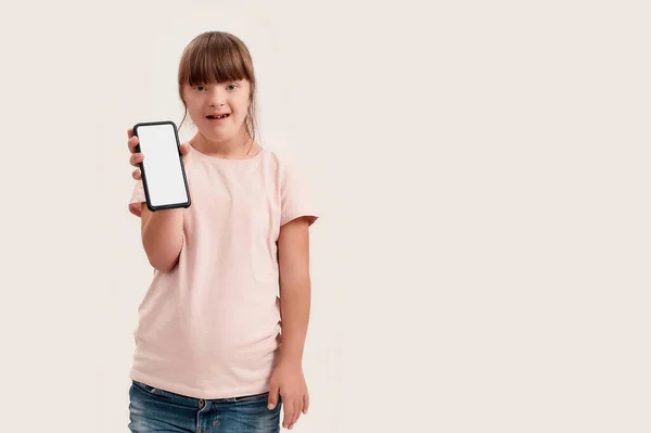 Retrato de chica discapacitada con síndrome de Down mirando a la cámara mientras sostiene el teléfono inteligente con pantalla en blanco, posando aislado sobre fondo blanco — Foto de Stock