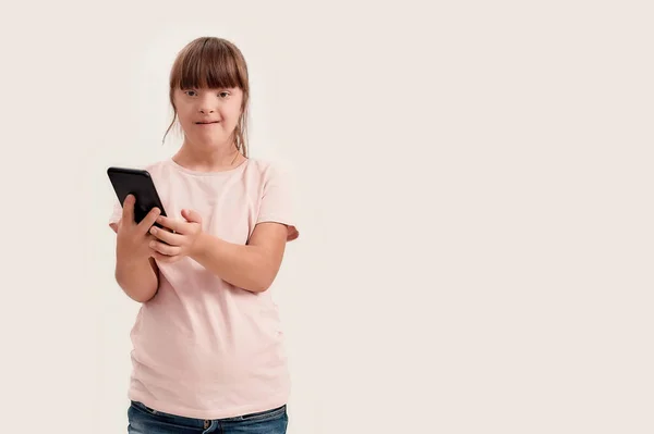 Retrato de chica discapacitada con síndrome de Down mirando a la cámara mientras usa el teléfono inteligente, de pie aislado sobre fondo blanco — Foto de Stock