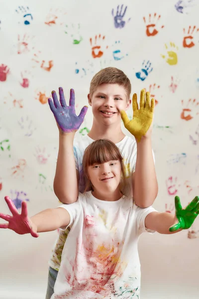 Bambini felici, ragazzo disabile e ragazza con la sindrome di Down sorridenti alla macchina fotografica, mostrando le mani dipinte con vernici colorate per le stampe a mano sul muro — Foto Stock
