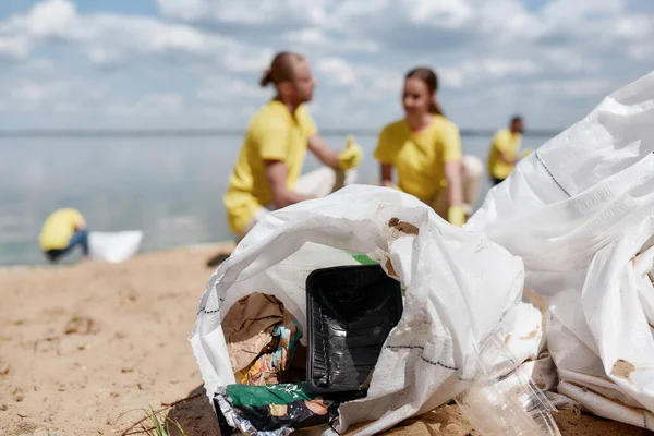 Bolsa de basura con residuos de plástico y otra basura de pie en la arena, dos jóvenes voluntarios en uniforme amarillo limpieza de la playa en el fondo, enfoque selectivo — Foto de Stock