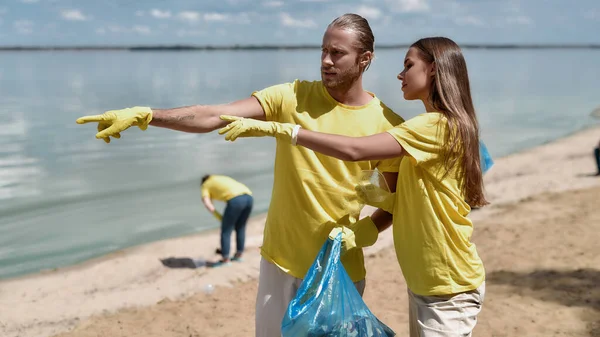 Dos jóvenes eco activistas, hombre y mujer sosteniendo la bolsa de basura y discutiendo algo mientras recogen residuos plásticos en la playa con un grupo de voluntarios — Foto de Stock