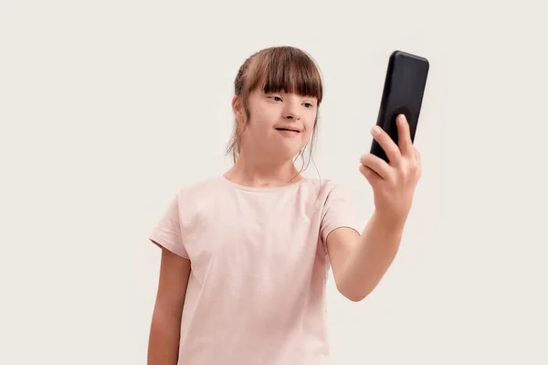 Portrait of disabled girl with Down syndrome using smartphone while taking selfie picture, standing isolated over white background — Stock Photo, Image