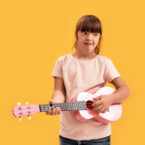 Menina deficiente alegre com síndrome de Down olhando para o lado enquanto joga ukulele, de pé isolado sobre fundo amarelo — Fotografia de Stock