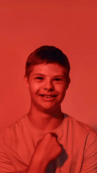 Retrato de adolescente feliz com deficiência menino com síndrome de Down sorrindo para a câmera, apontando para si mesmo enquanto posando isolado sobre fundo de luz vermelha — Fotografia de Stock