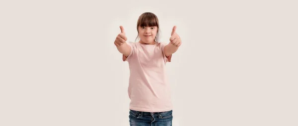 Portrait of disabled girl with Down syndrome smiling at camera, showing thumbs up with both hands while standing isolated over white background — Stock Photo, Image