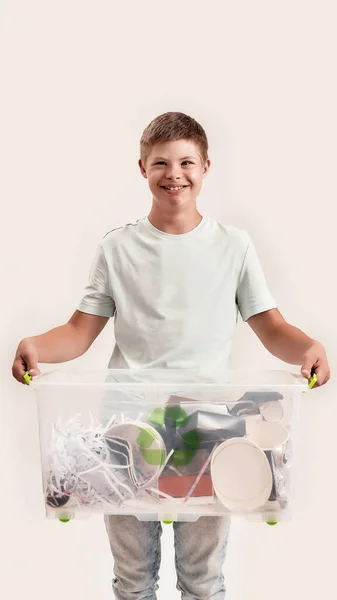 Cheerful disabled boy with Down syndrome smiling at camera while holding a recycling bin full of paper waste, standing isolated over white background — Stock Photo, Image