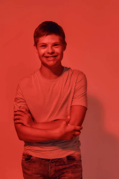 Portrét šťastného dospívajícího zdravotně postiženého chlapce s Downovým syndromem usmívajícího se do kamery, zatímco stojí s rukama zkříženýma na červeném pozadí — Stock fotografie