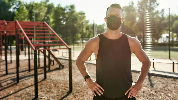 Ενεργός νεαρός φορώντας μάσκα, κοιτάζοντας την κάμερα, στέκεται σε εξωτερικούς χώρους, ενώ εργάζονται έξω στο υπαίθριο γυμναστήριο — Φωτογραφία Αρχείου