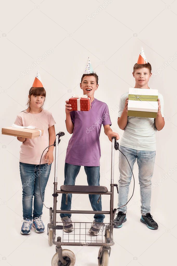 Full length shot of three disabled children wearing birthday caps smiling at camera and holding presents while celebrating birthday together isolated over white background