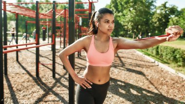 Güneşli bir günde açık hava spor salonunda lastik bantla antrenman yapan odaklanmış genç bir kadının portresi.
