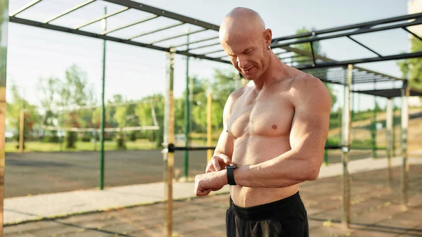 Sportive muscular half naked man checking results using his smart watch while working out at outdoor gym on a sunny day — Stok Foto