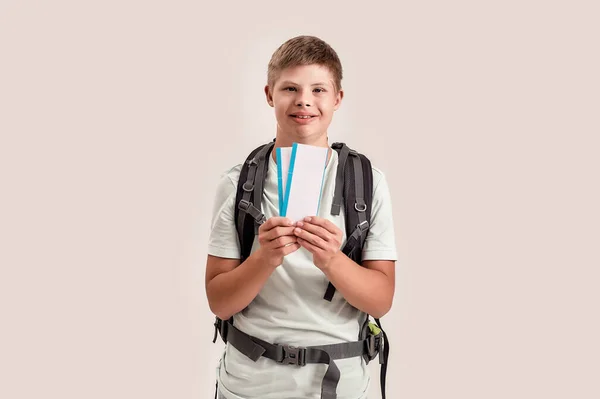 Niño discapacitado feliz con síndrome de Down usando mochila sonriendo mientras sostiene boletos aéreos, de pie aislado sobre fondo blanco — Foto de Stock