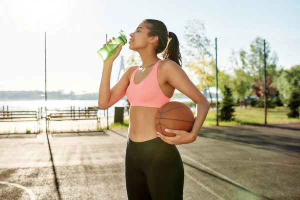 Attractive young female basketball player drinking water from bottle, holding basketball while standing outdoors on a sunny day