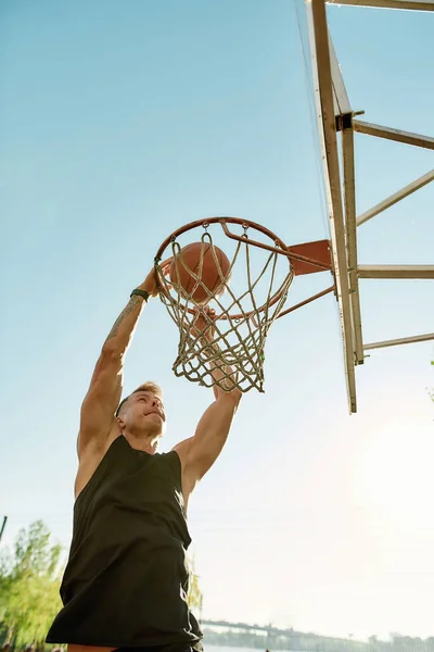 Skilled sportive guy throwing ball into basket, while playing basketball outdoors