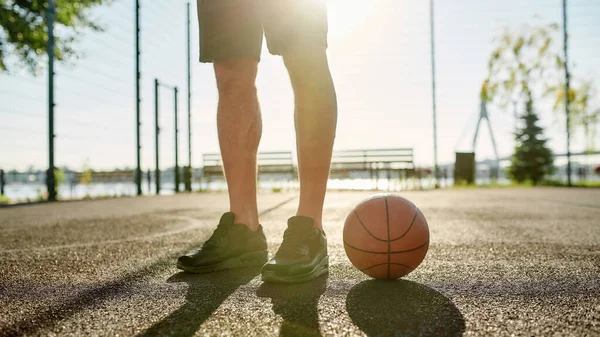 Primo piano delle gambe del giovane giocatore di basket in abbigliamento sportivo in piedi sul campo con il basket in una giornata di sole — Foto Stock