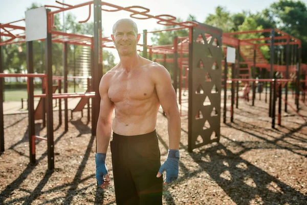 Mature athletic man with naked torso and boxing bandage on his fists smiling at camera, standing in boxing pose while having workout at street gym yard — Stok Foto