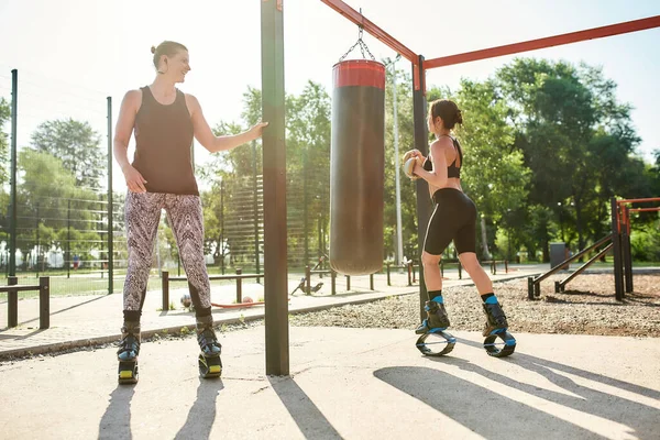 Tiro de longitud completa de dos mujeres deportivas en kangoo zapatos de salto de entrenamiento con saco de boxeo, tener entrenamiento en el patio del gimnasio de la calle —  Fotos de Stock