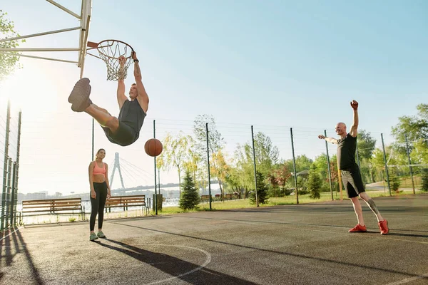 Skilled guy throwing ball into basket, while playing basketball. Full length shot of active young people spending time outdoors on playground — Fotografia de Stock
