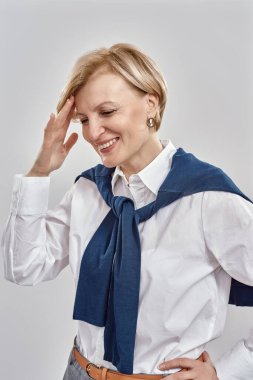 Kafkasyalı zarif bir orta yaşlı kadının portresi iş elbisesi giymiş, gülümseyerek kafasına dokunuyor, gri arka planda soyutlanmış bir şekilde poz veriyor.