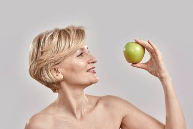 Güzel orta yaşlı kadın yeşil elmaya bakıyor, gri arka planda soyutlanmış poz veriyor.