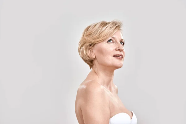 Retrato de una hermosa mujer de mediana edad en sujetador blanco mirando hacia arriba, posando aislado sobre fondo gris. Concepto de belleza —  Fotos de Stock