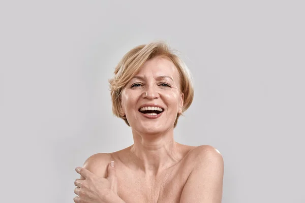 Retrato de bela mulher de meia-idade rindo da câmera, tocando sua pele, posando isolada contra fundo cinza. Beleza, conceito de cuidado da pele — Fotografia de Stock