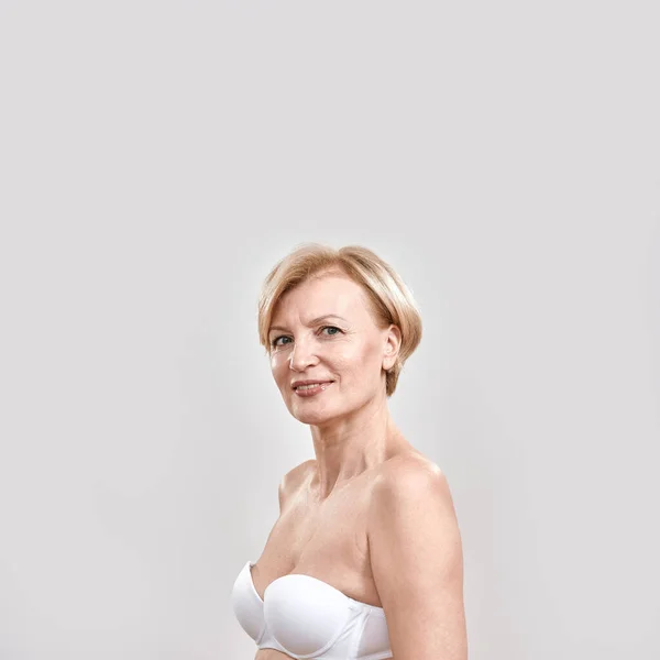 Retrato de una alegre y hermosa mujer de mediana edad en sujetador blanco mirando a la cámara, posando aislada sobre un fondo gris — Foto de Stock