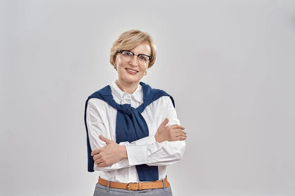 Портрет элегантной белой женщины средних лет в деловой одежде и очках, улыбающейся в камеру во время позирования, стоящей со скрещенными руками на сером фоне Стоковая Картинка