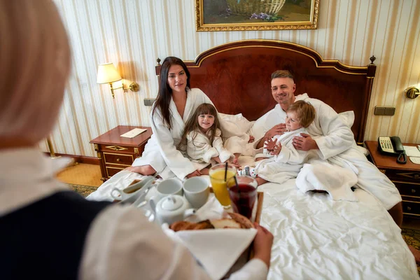Щасливого життя. Кавказькі батьки і двоє дітей в білому халаті посміхаються, чекаючи на сніданок, доставлений офіціанткою в розкішному готельному номері. Сім'я, подорожі, курорт, ідея обслуговування кімнат — стокове фото