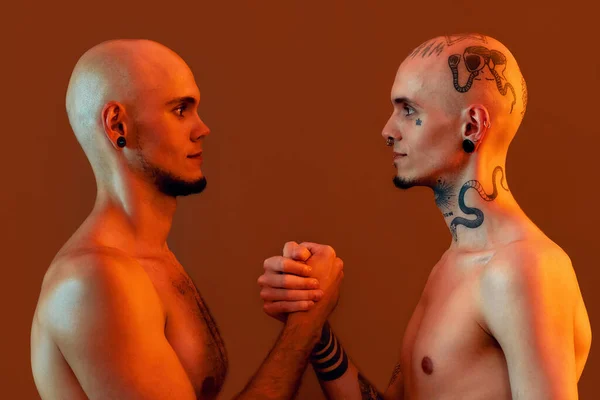 Ritratto di giovani fratelli gemelli con tatuaggi e piercing che si guardano, si tengono per mano, in piedi faccia a faccia isolati su sfondo arancione — Foto Stock