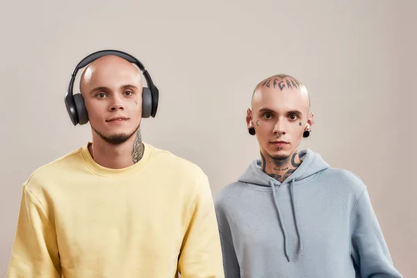 Dois jovens irmãos gêmeos caucasianos em desgaste casual com tatuagens e piercings ouvindo música em fones de ouvido e fones de ouvido sem fio, de pé isolados em fundo bege — Fotografia de Stock