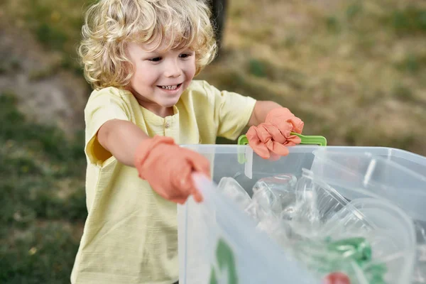 Recycling and Kids. Cute little boy wearing rubber gloves holding recycle bin and smiling while collecting plastic waste in forest or park with parents Royalty Free Stock Photos