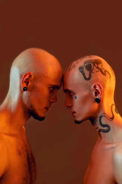 Πορτρέτο των νέων δίδυμων αδελφών με τατουάζ και σκουλαρίκια στέκεται κεφάλι με κεφάλι, κοιτάζοντας ο ένας τον άλλο απομονωμένο πάνω από πορτοκαλί φόντο — Φωτογραφία Αρχείου