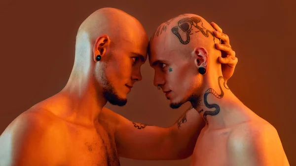 Πορτρέτο των νέων δίδυμων αδελφών με τατουάζ και σκουλαρίκια στέκεται κεφάλι με κεφάλι, κοιτάζοντας ο ένας τον άλλο απομονωμένο πάνω από πορτοκαλί φόντο — Φωτογραφία Αρχείου