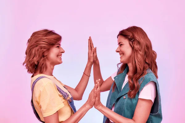매력적 인 두 소녀가, 쌍동이 자매들이 손바닥에 결합하여, 핑크 색 배경 위로 따로 모여 앉아 서로 미소짓고 있는 모습 — 스톡 사진