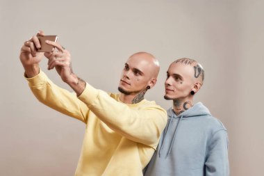 En iyi arkadaşla fotoğraf. Gündelik giyinmiş iki beyaz erkek kardeş bej arka planda tek başına dururken cep telefonunda selfie çekiyorlar.
