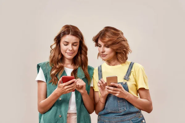 2人の魅力的な若い女の子,一緒にポーズを取りながら、スマートフォンアプリを使用してカジュアルな服装の双子の姉妹,光の背景に孤立立って — ストック写真
