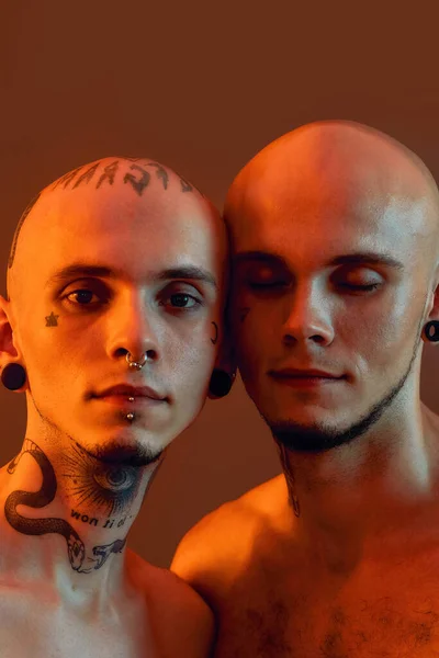 Close up retrato de jovens meio nus irmãos gêmeos com tatuagens e piercings posando juntos isolado sobre fundo laranja — Fotografia de Stock
