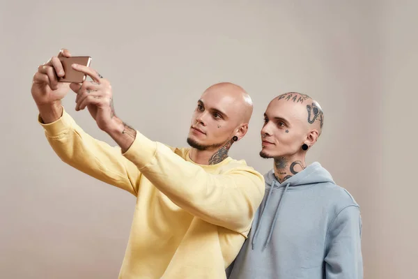Foto med bästa vän. Två unga kaukasiska tvillingbröder i casual wear tar selfie på mobiltelefon samtidigt står tillsammans isolerade över beige bakgrund — Stockfoto
