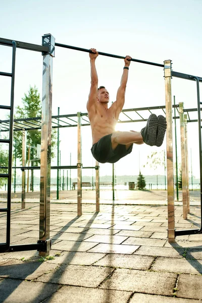 Joven atlético colgando de las barras en el gimnasio de calistenia al aire  libre sonriendo