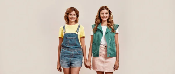 Retrato de dos chicas jóvenes atractivas, hermanas gemelas en ropa casual sonriendo a la cámara, cogidas de la mano, posando juntas, de pie aisladas sobre un fondo claro — Foto de Stock